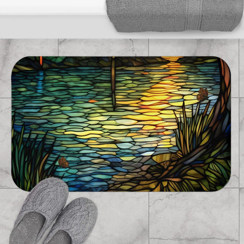 Puede incluir: Una alfombra de ba&ntilde;o de vidrio tintado con un dise&ntilde;o de un lago con una puesta de sol reflejada en el agua. La alfombra tiene un borde marr&oacute;n oscuro y una variedad de colores, incluyendo azul, verde, amarillo y naranja.