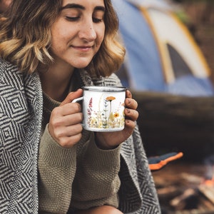 May include: A white enamel mug featuring a colorful watercolor illustration of various wildflowers including orange poppies, yellow blossoms, and tall purple stalks, finished with a dark metallic rim and a matching handle on the side.