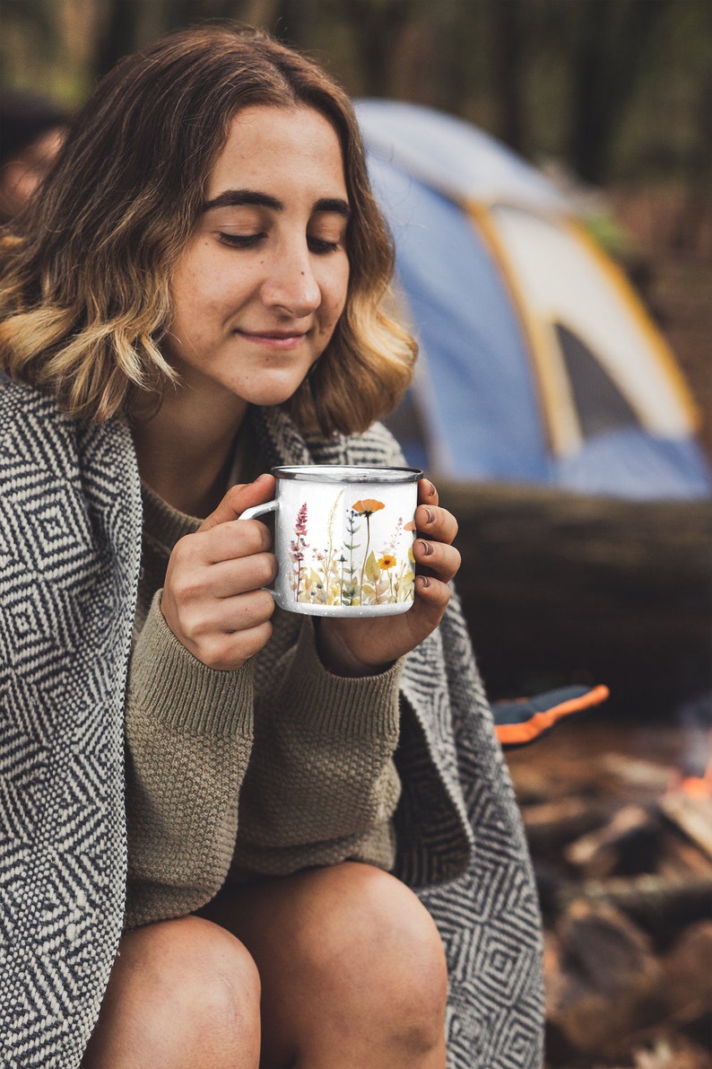 May include: A white enamel mug featuring a colorful watercolor illustration of various wildflowers including orange poppies, yellow blossoms, and tall purple stalks, finished with a dark metallic rim and a matching handle on the side.