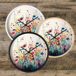 May include: Three round wall clocks featuring a watercolor illustration of a wildflower meadow with red, pink, blue, and yellow flowers, green stems, and a dragonfly. The clocks have black hands and frames in white, light wood, and black colors.