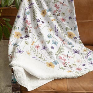 Op de afbeelding: Een wit deken met een bloemenpatroon met delicate bloemen, bladeren en libellen in tinten roze, geel, blauw en paars. Het deken heeft een zachte, witte sherpa voering.