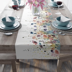 May include: A floral table runner with a watercolor design featuring pink, blue, and yellow flowers. The runner is displayed on a wooden table set with teal-colored bowls, plates, and wine glasses. A vase of pink lilies is in the center.