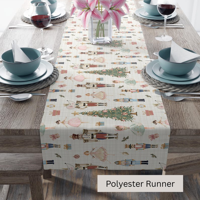 May include: A cream-colored polyester table runner with a repeating pattern of Christmas trees, nutcrackers, and ballerinas. The runner is set on a wooden table with place settings and a vase of pink flowers. The text "Polyester Runner" is visible.