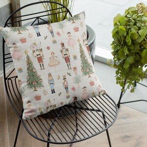 May include: A decorative square pillow with a festive Nutcracker theme. The pillow features a repeating pattern of Nutcracker soldiers, ballerinas, Christmas trees, and gift boxes in soft pastel colors on a cream background. The pillow is placed on a black metal chair.