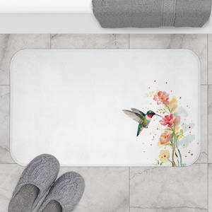 May include: White rectangular bath mat featuring a watercolor hummingbird and floral design in shades of pink, yellow, and green. The mat has rounded corners and is placed on a tiled floor. Gray slippers are in the foreground.