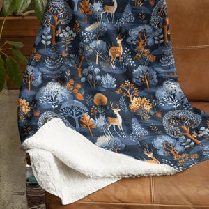 Puede incluir: Una manta suave y mullida con fondo azul y un patrón repetitivo de árboles estilizados, ciervos y otros elementos del bosque en tonos naranja, marrón y blanco.