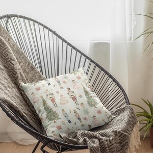 May include: A decorative pillow with a festive pattern featuring nutcrackers, Christmas trees, and other holiday elements. The pillow is placed on a black wire chair with a patterned throw blanket. The scene is set in front of a window.