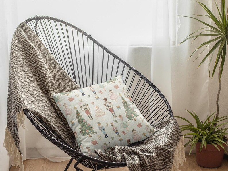 May include: A decorative pillow with a festive pattern featuring nutcrackers, Christmas trees, and other holiday elements. The pillow is placed on a black wire chair with a patterned throw blanket. The scene is set in front of a window.