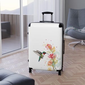 May include: A white hard-shell rolling suitcase featuring a watercolor illustration of a hummingbird in flight near a cluster of pink and yellow flowers. The luggage has a black retractable handle, black side trim, and four black spinner wheels.