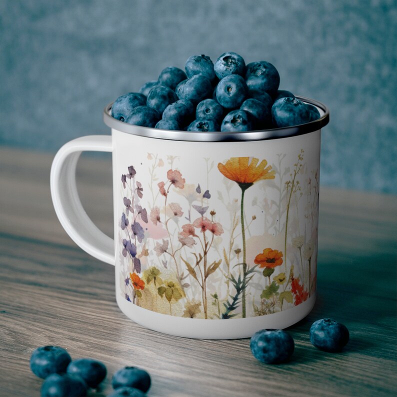 May include: A white enamel mug with a silver rim, decorated with a watercolor-style print of various wildflowers in shades of orange, pink, and purple. The mug is filled to the brim with fresh, dark blue blueberries, with several scattered nearby.