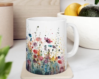 May include: A white ceramic mug featuring a colorful watercolor print of wildflowers in shades of red, blue, yellow, and purple. The design covers the exterior surface with delicate stems and splattered paint effects, complemented by a handle.