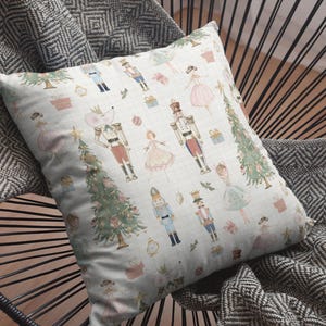 May include: A decorative pillow with a light blue background featuring illustrations of nutcrackers, ballerinas, Christmas trees, and gifts. The pillow is placed on a black chair with a gray and white patterned blanket.