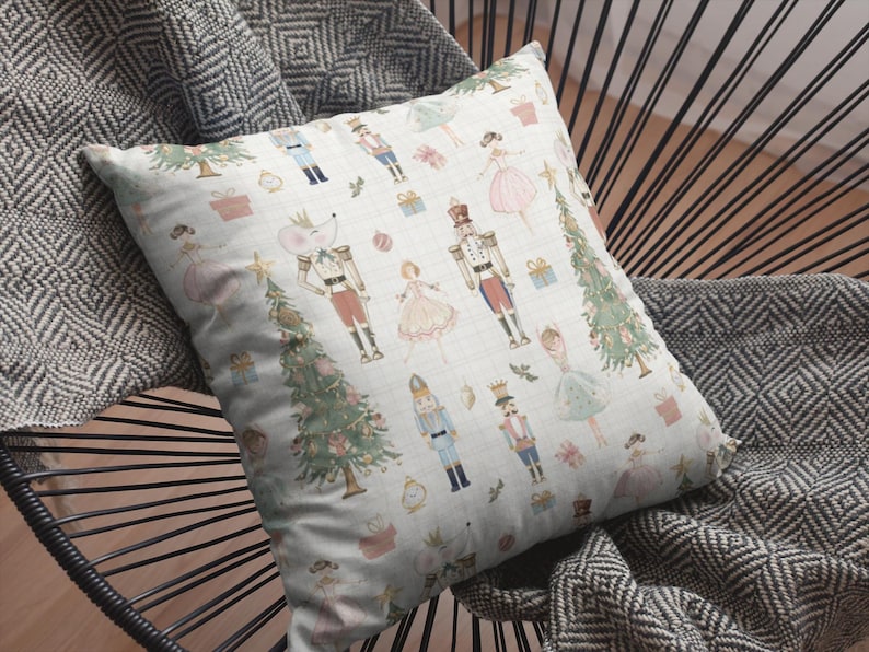 May include: A decorative pillow with a light blue background featuring illustrations of nutcrackers, ballerinas, Christmas trees, and gifts. The pillow is placed on a black chair with a gray and white patterned blanket.