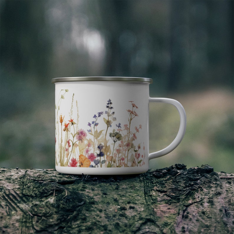 May include: A white enamel mug featuring a colorful watercolor-style print of various wildflowers, including small blue, pink, orange, and yellow blossoms with green stems. The mug has a smooth white finish, a curved handle, and a metallic silver rim.