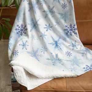 May include: A white fleece throw blanket with a blue watercolor snowflake pattern. The blanket is folded and draped over a brown leather couch.