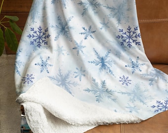 Könnte beinhalten: Eine weiße Fleece-Decke mit einem blauen Aquarell-Schneeflockenmuster. Die Decke ist gefaltet und über ein braunes Ledersofa gelegt.
