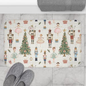 May include: Rectangular bath mat with a whimsical Nutcracker theme. The mat features illustrations of Nutcracker soldiers, ballerinas, Christmas trees, and gift boxes on a cream-colored background with a grid pattern. Gray slippers are at the bottom.