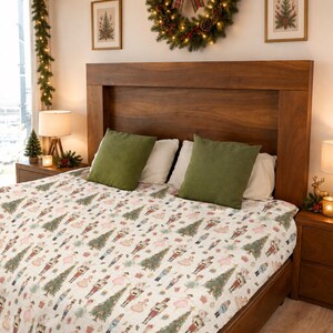 May include: A white bedspread featuring a repeating pattern of nutcracker figures, Christmas trees, and small gift boxes. The bedding is placed on a wooden bed frame with two olive green square pillows and two white rectangular pillows.
