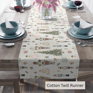 May include: A cotton twill table runner with a repeating pattern of nutcrackers, Christmas trees, and ballerina figures in pastel colors. The runner is displayed on a wooden table set for a meal, with plates, bowls, and wine glasses.