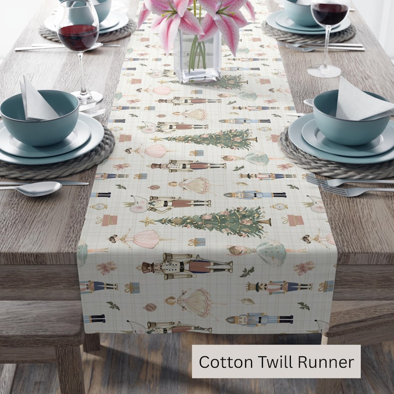 May include: A cotton twill table runner with a repeating pattern of nutcrackers, Christmas trees, and ballerina figures in pastel colors. The runner is displayed on a wooden table set for a meal, with plates, bowls, and wine glasses.