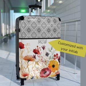 May include: A rolling suitcase featuring a black and white geometric pattern on the top section and a white base with a watercolor floral print of red, orange, and yellow flowers. The suitcase displays the initials MSW and a yellow arrow graphic.