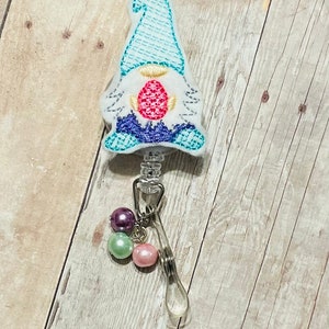 May include: A badge reel featuring a gnome design with a light blue hat and an Easter egg. The gnome is white with blue and purple detailing. The reel has a clear clip and a charm with purple, green, and pink beads.