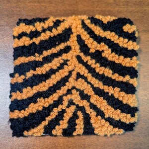 May include: Square, textured textile with a repeating pattern of black and orange stripes. The stripes are arranged in a zig-zag design, creating a visually interesting and tactile surface. The textile appears to be made of yarn or a similar material.