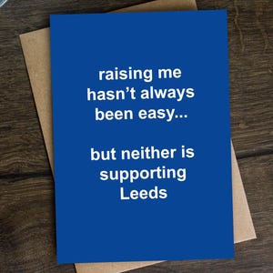May include: A blue greeting card with the text "raising me hasn't always been easy... but neither is supporting Leeds" is on a brown envelope. A silver pen rests on a dark wood surface, with a plant in the upper left corner.