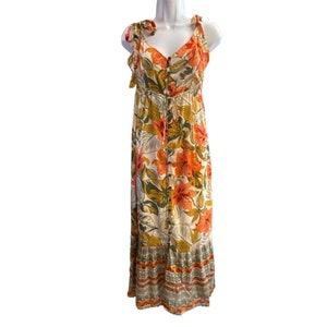 May include: A white maxi dress with a floral print in shades of orange, green, and yellow. The dress has a V-neckline, adjustable straps, and a button-up front. The hemline is tiered and features a colorful trim.