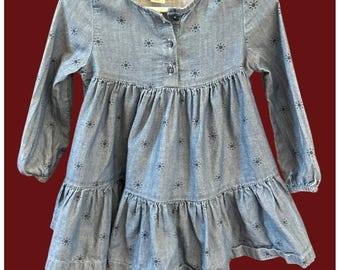 May include: A light blue denim dress with long sleeves and a tiered skirt. The dress has a small floral pattern and a button-down front. The dress is hanging on a white hanger.