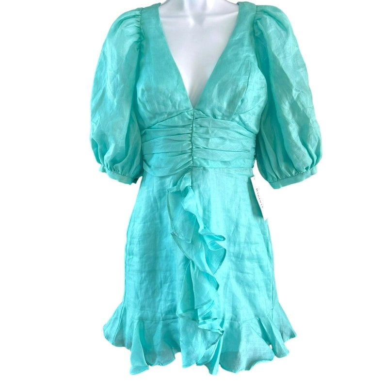 May include: A turquoise blue linen mini dress with a ruffled hem and puffed sleeves. The dress has a deep V-neckline and a fitted bodice.
