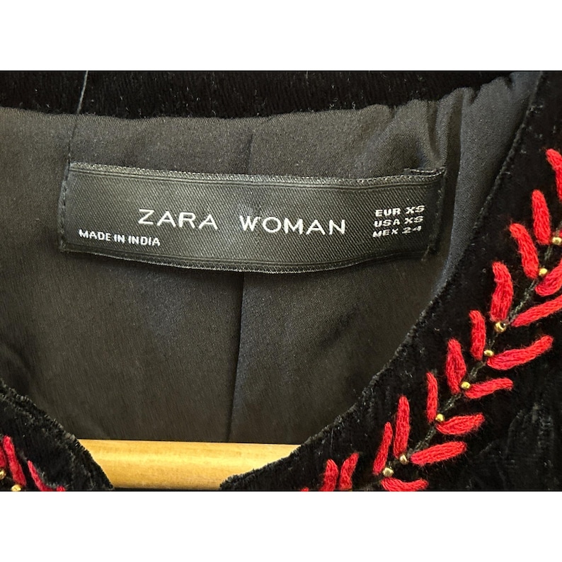 May include: Close-up of a black velvet jacket with red embroidered leaf-like designs and gold bead accents. The label reads "ZARA WOMAN" and "MADE IN INDIA". The jacket is size EUR XS, USA XS, and MEX 24.