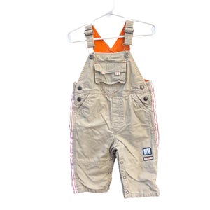 May include: Beige overalls featuring orange shoulder straps, a front chest pocket with a flap, and red and white striped side panels. The garment includes metal buttons, a small square patch on the leg, and a rectangular patch labeled PLAYER 1.
