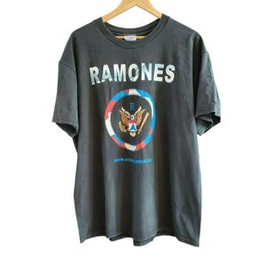May include: A black t-shirt with the word "RAMONES" printed in white on the front. The shirt features a circular graphic of an eagle with a baseball bat and the text "www.officialramones.com".
