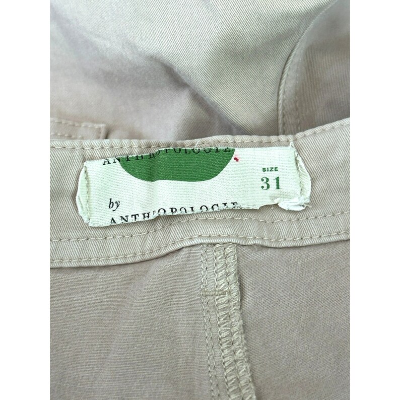 May include: A beige pair of pants with a label that reads "by Anthropologie" and "size 31".