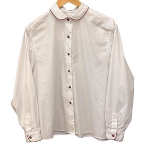 May include: A long-sleeved white button-down shirt featuring a rounded Peter Pan collar with a thin dark red trim. The shirt includes a front button placket with matching dark red buttons and buttoned cuffs, each accented with the same dark red trim.