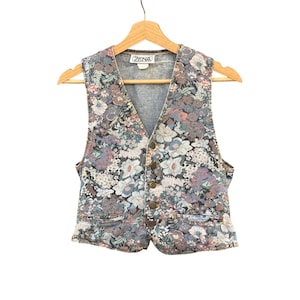 Pode incluir: Um colete sem mangas com um padrão floral denso em tons de azul, rosa e creme. A peça tem um decote em V, um fecho frontal com quatro botões metálicos e dois pequenos bolsos embutidos na cintura. Uma etiqueta da marca ZENA é visível.