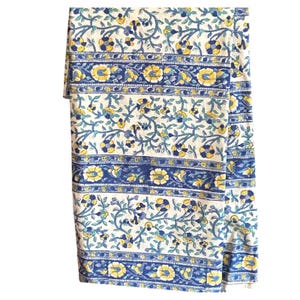May include: A folded, cream-colored textile with a repeating floral pattern in blue and yellow. The design includes horizontal bands of solid blue and floral borders, creating a decorative and patterned look. The fabric appears to be cotton or linen.