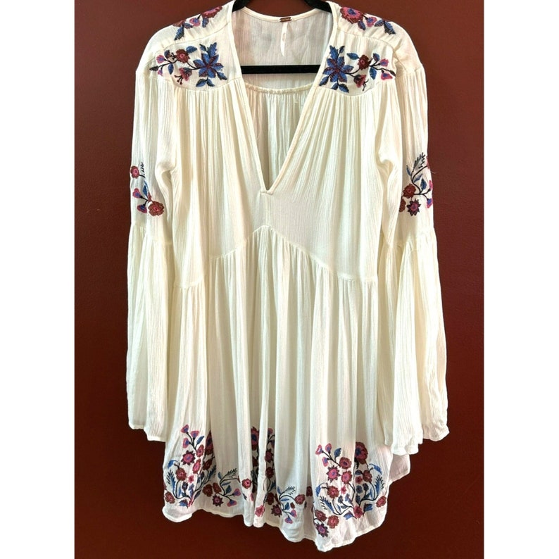 May include: A white, long-sleeved, embroidered tunic with a V-neckline and bell sleeves. The embroidery features floral designs in shades of blue, pink, and red.