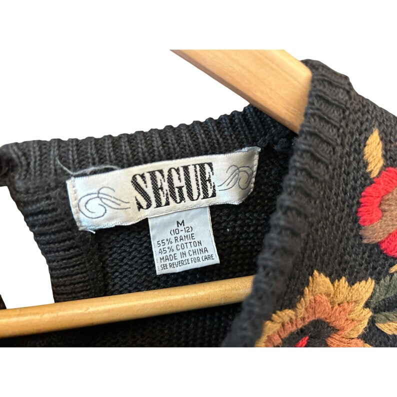 May include: Close-up of a black sweater with floral embroidery in shades of red, yellow, and brown. The label reads "SEGUE" and includes size and material information. The sweater is made of 55% ramie and 45% cotton.