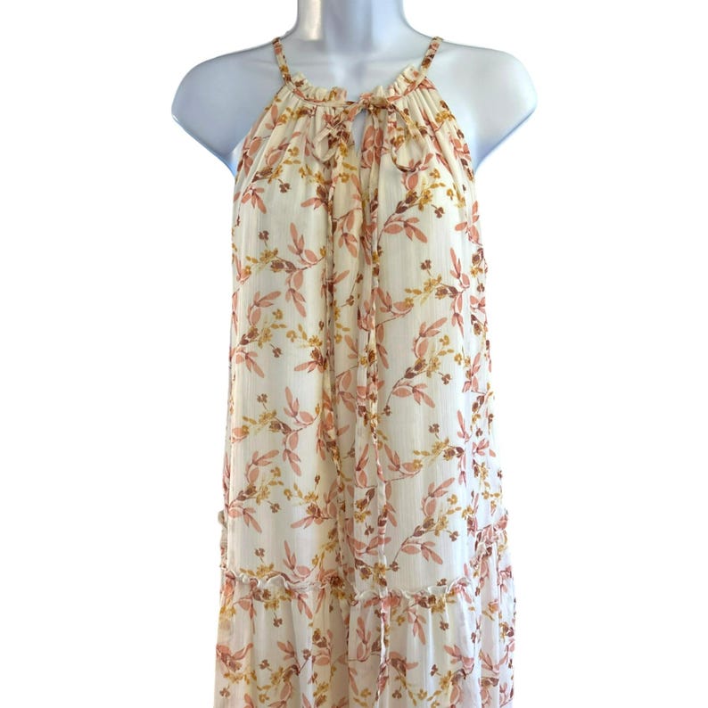 Pode incluir: Um vestido maxi branco com estampa floral com flores rosa e amarelas. O vestido tem um decote halter com um la&ccedil;o nas costas e uma saia em camadas.