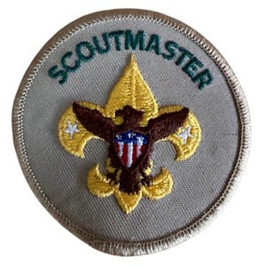 May include: Embroidered Scoutmaster patch featuring a gold fleur-de-lis, a brown eagle, and the text "SCOUTMASTER" in green. The patch has a grey background and a stitched border.