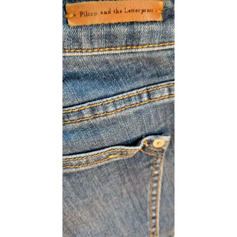 May include: Close-up of a pair of blue denim jeans with a brown leather tag that reads "Pilcro and the Letterpress".