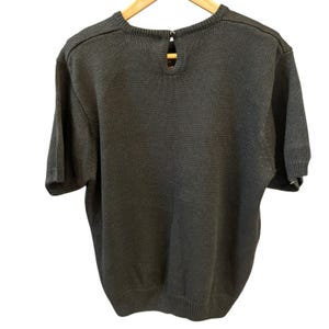 May include: A dark gray short-sleeve sweater with a round neckline and a keyhole detail at the back. The sweater is made of a knitted fabric and hangs on a wooden hanger. The back of the sweater is visible.