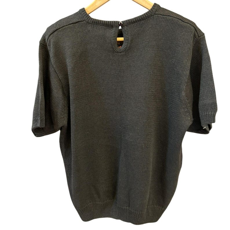 May include: A dark gray short-sleeve sweater with a round neckline and a keyhole detail at the back. The sweater is made of a knitted fabric and hangs on a wooden hanger. The back of the sweater is visible.