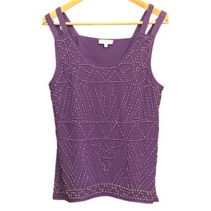 Puede incluir: Una camiseta sin mangas morada con cuello redondo y tirantes dobles. La parte superior presenta un patrón geométrico de costuras blancas en la parte delantera. La camiseta sin mangas está colgada en una percha de madera.