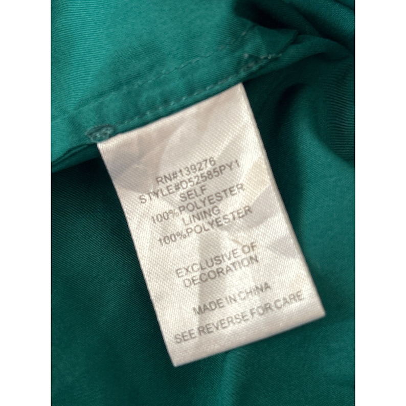 May include: White tag with black text detailing the fabric content and care instructions for a garment. The tag reads "RN#139276 STYLE#052585PY1 SELF 100% POLYESTER LINING 100% POLYESTER EXCLUSIVE OF DECORATION MADE IN CHINA SEE REVERSE FOR CARE".