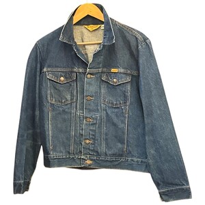 May include: A dark blue denim jacket featuring a pointed collar, long sleeves, and a front button closure. The jacket includes two chest pockets with buttoned flaps and a yellow brand label on the interior neck and above the left pocket.