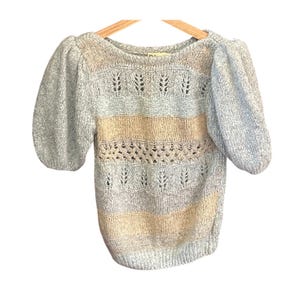 May include: A knitted jumper in shades of light blue, beige, and grey, with short, puffed sleeves. The design includes horizontal stripes and openwork patterns. The garment is displayed on a wooden hanger.
