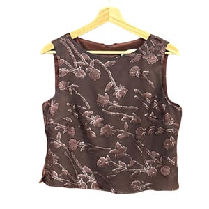 May include: A sleeveless brown top with a floral pattern in a lighter shade of brown. The top is hanging on a wooden hanger against a white background. The floral design covers the entire front of the top.